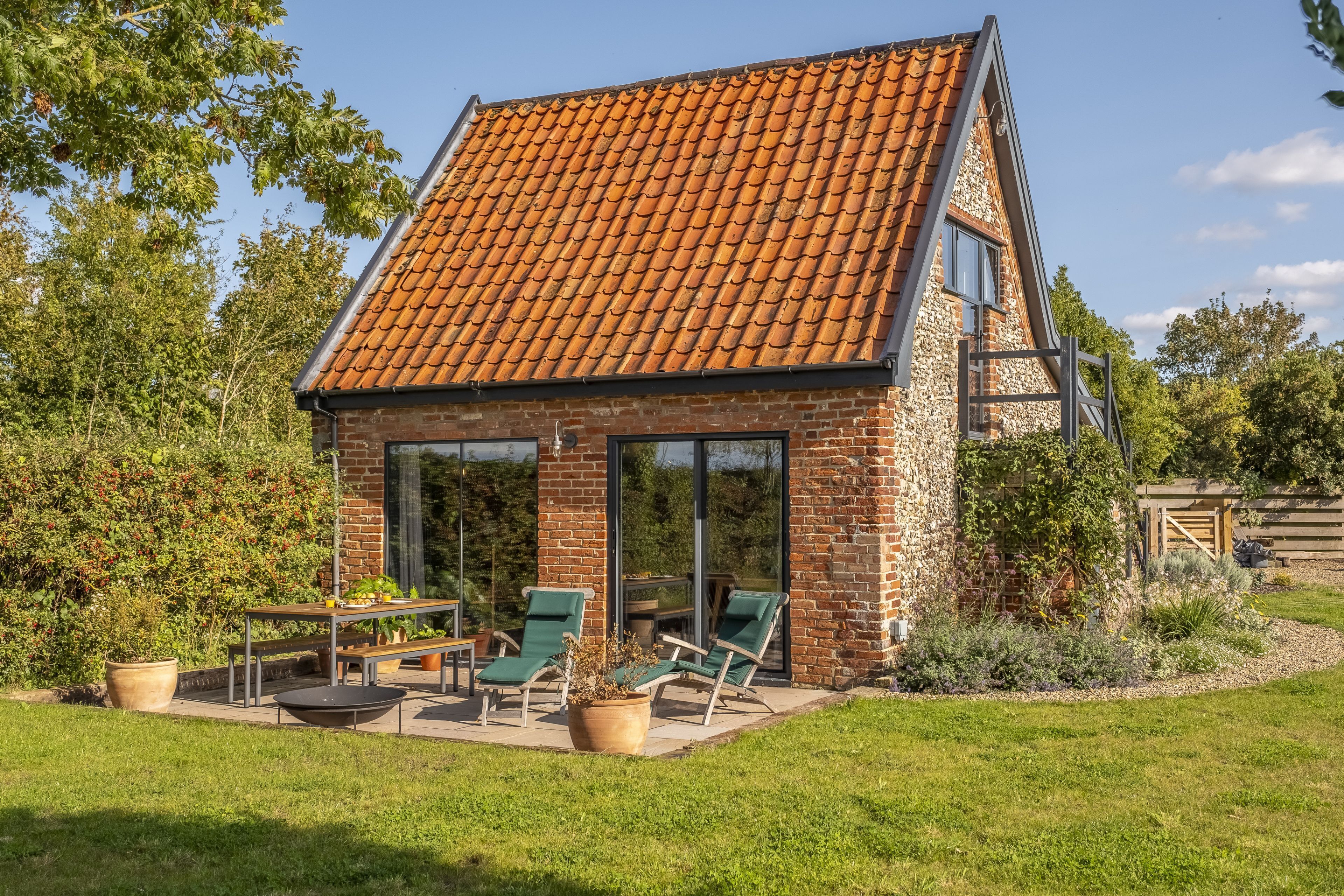 Une petite maison en briques avec un toit en pente, de grandes portes vitrées et une terrasse aménagée avec du mobilier d’extérieur.