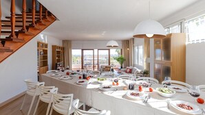 Un comedor amplio con una mesa larga dispuesta para una comida, rodeado de sillas blancas, y una escalera a un lado.