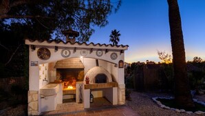 Horno de ladrillo exterior con fachada de piedra y azulejos decorativos, rodeado por un sendero de grava y palmeras.