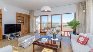 Amplia sala de estar con una televisión de pantalla plana grande, una mesa de centro de vidrio y un sofá con un cojín rojo. Hay un balcón con vistas a la ciudad y a las montañas.