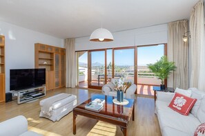 Ein geräumiges Wohnzimmer mit einem großen Flachbildfernseher, einem Glastisch und einem Sofa mit einem roten Kissen. Ein Balkon bietet Blick auf die Stadt und die Berge.