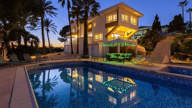 Una casa moderna de dos pisos con piscina, rodeada de palmeras y mobiliario de exterior.
