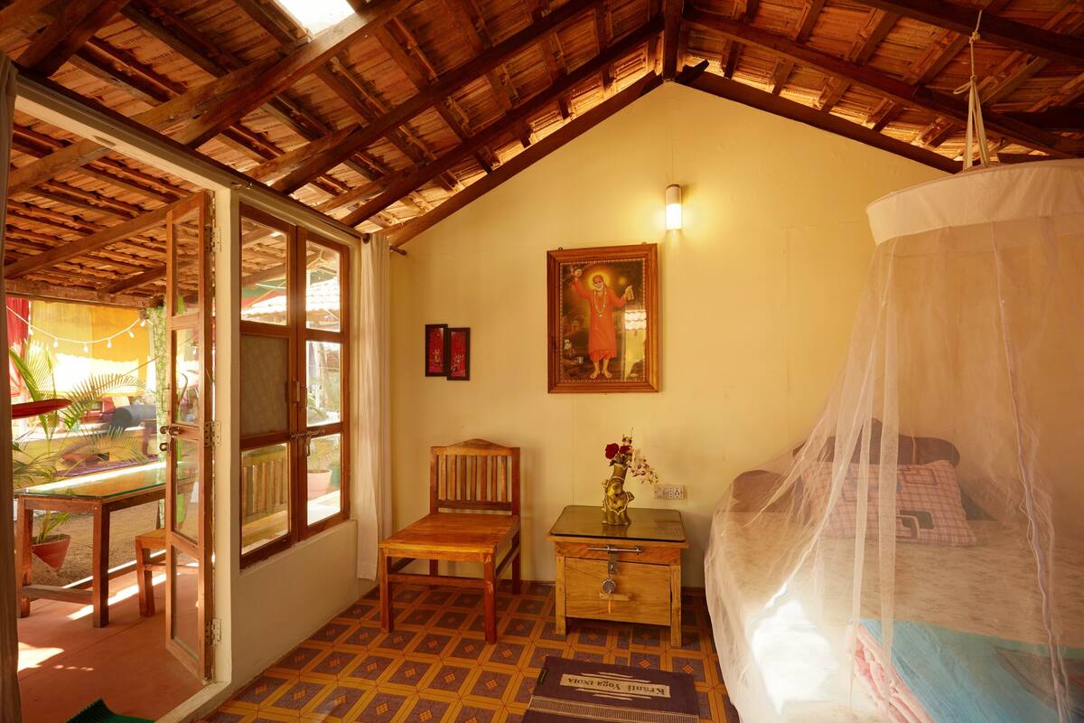 Ein gemütliches Zimmer mit Holzbalkendecke, einem Bett mit Moskitonetz, einem Holzstuhl, einem kleinen Tisch und einem an der Wand hängenden Bild.