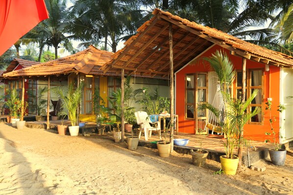 A row of rustic, single-story buildings with tiled roofs, surrounded by potted plants and palm trees. - Kranti Yoga Tradition (Canacona)