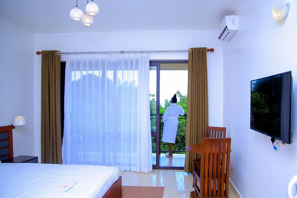 A hotel room with a bed, a TV, a balcony with a person in a robe, and curtains.