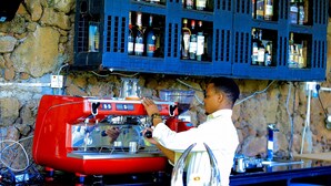 Un barista prépare une boisson sur une machine à expresso rouge, avec des étagères remplies de bouteilles de spiritueux au-dessus.