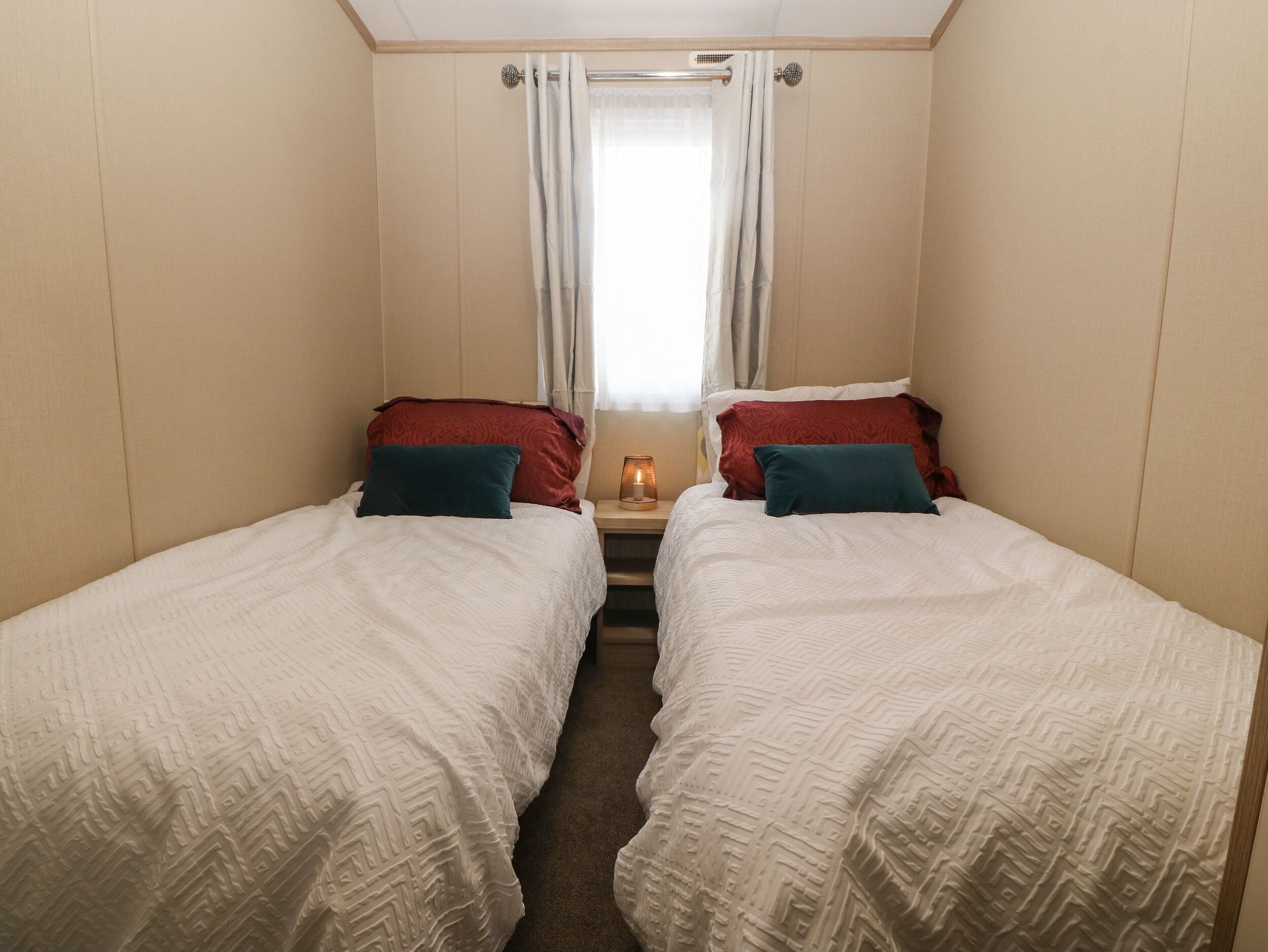 Quarto pequeno e bem iluminado com duas camas individuais, cada uma com colcha branca e almofadas vermelhas e azul-esverdeadas. Uma pequena mesa com abajur fica entre as camas.