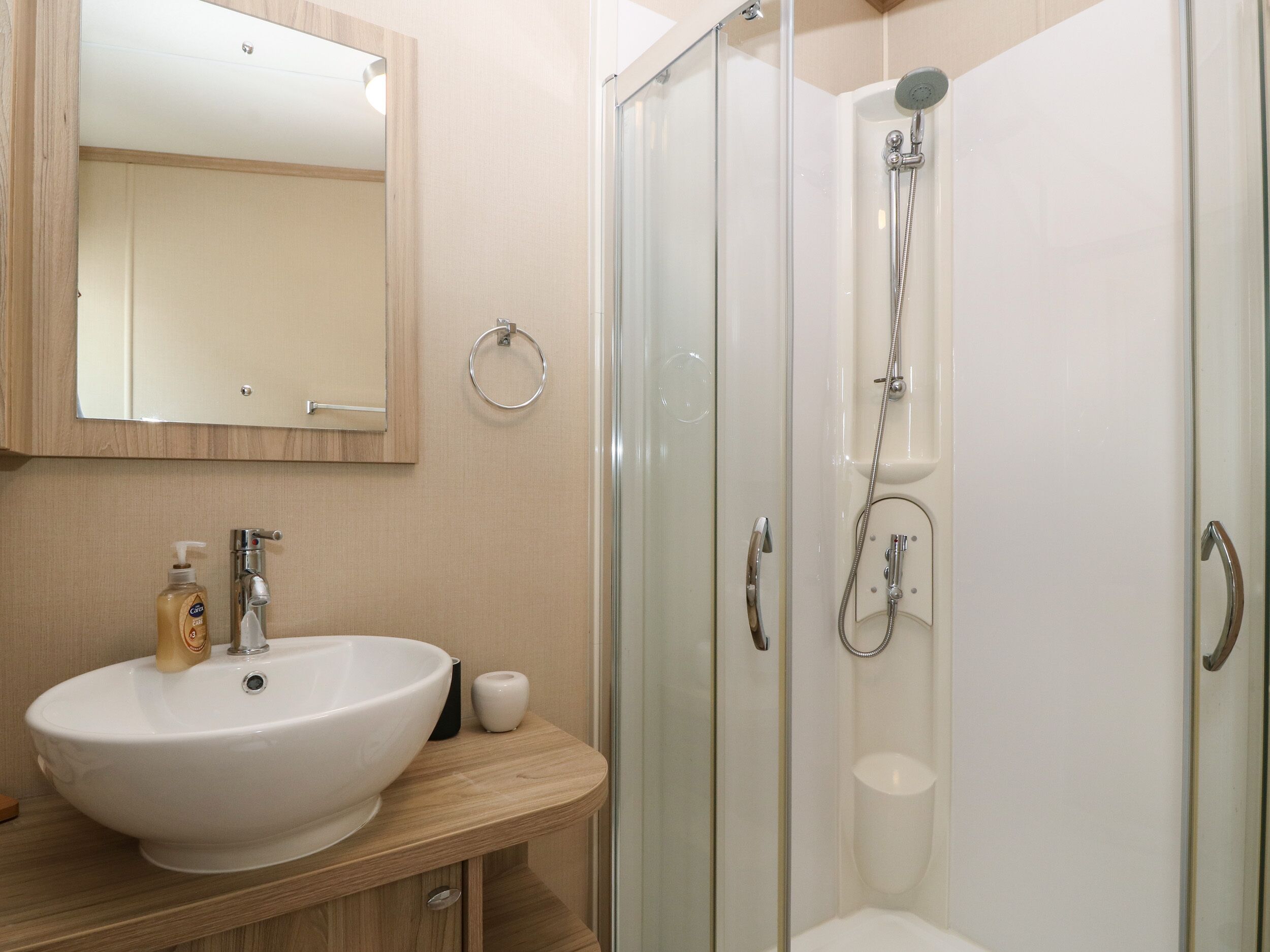 Een badkamer met een witte wastafel, een spiegel, een douchecabine van glas en een houten wastafelmeubel.