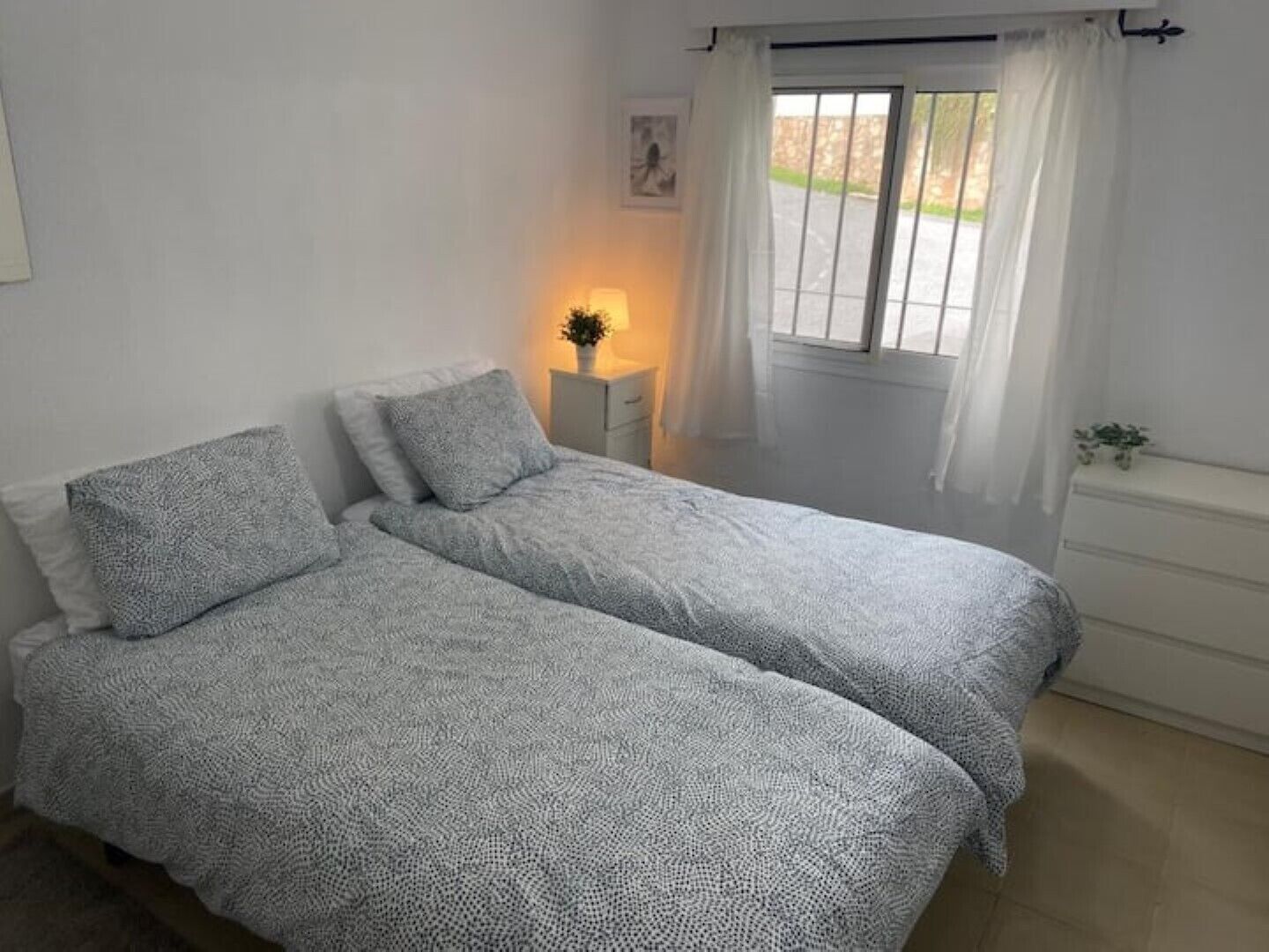 Een slaapkamer met twee bedden, een nachtkastje met een lamp, een raam met gordijnen en een kleine plant op de kaptafel.