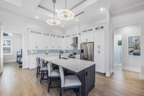 A modern kitchen with white cabinetry, a large island with bar stools, stainless steel appliances, and pendant lighting. - 2023 Custom Built Inland Home | Pool & Spa (Marco Island)