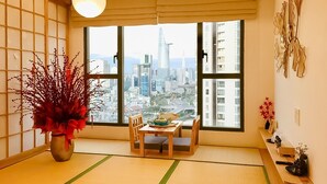 Una habitació tradicional japonesa amb paviment de tatami, una taula baixa amb coixins, un vas amb flors vermelles i una vista d'un paisatge urbà a través de grans finestres.
