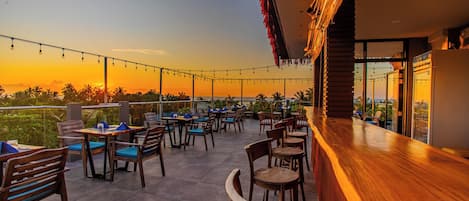 An outdoor seating area with tables and chairs, a wooden bar, and a view of the sunset over the ocean.