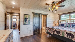 Une chambre de style cabane avec un plafond en bois, un lit recouvert d’une courtepointe à motifs d’ours, un ventilateur de plafond et une grande fenêtre.
