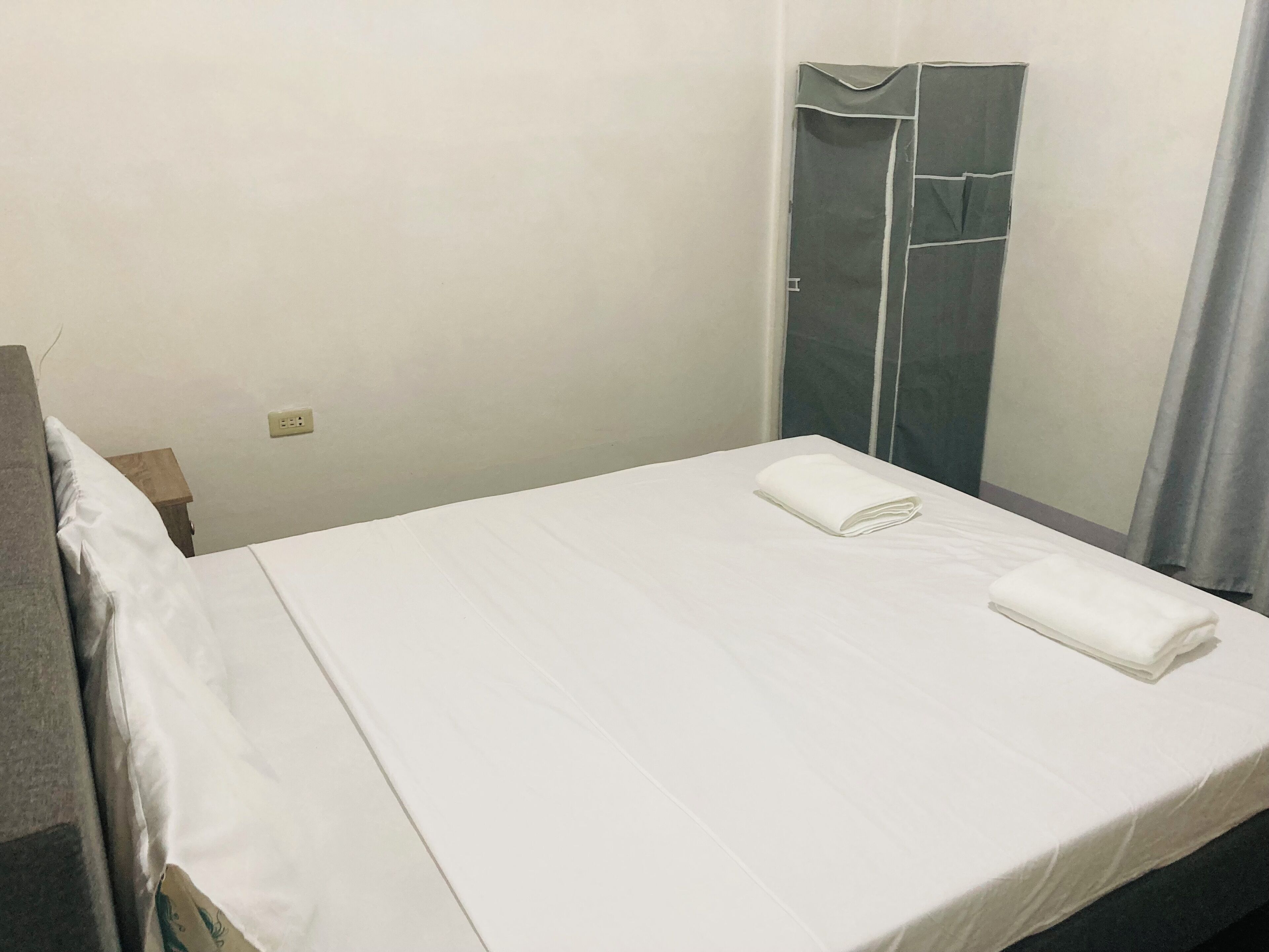 A neatly made bed with white linens and two folded towels, a wooden nightstand with a lamp, and a grey wardrobe with a mirror.
