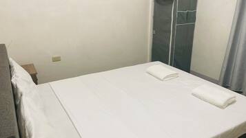 A neatly made bed with white linens and two folded towels, a wooden nightstand with a lamp, and a grey wardrobe with a mirror.