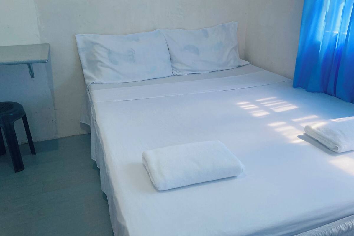 A neatly made bed with white linens and a folded towel, a small stool, and a window with blue curtains.