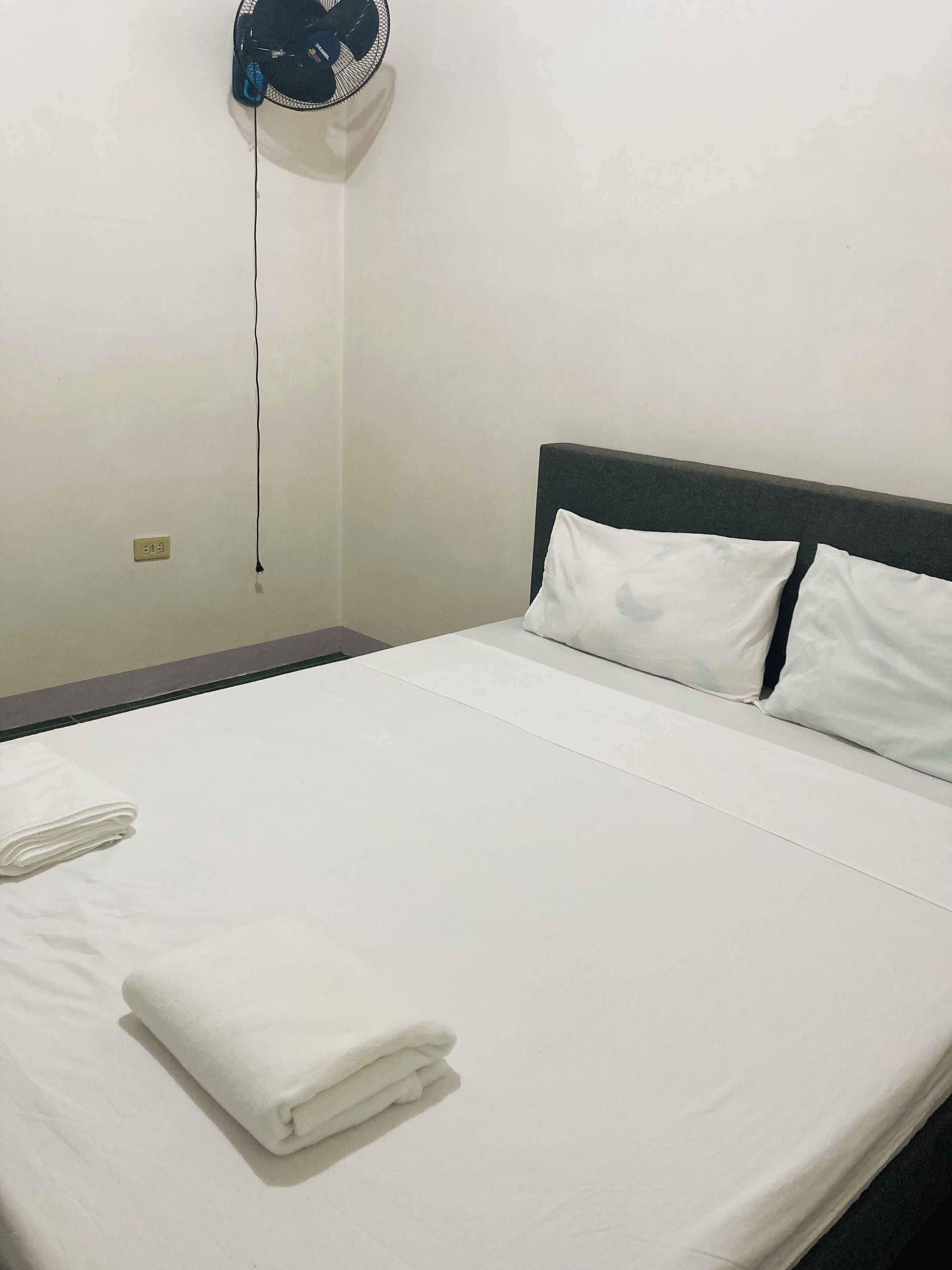 A neatly made bed with white linens and pillows, a folded white towel, and a wall-mounted fan.