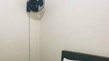 Uma cama bem arrumada com lençóis e almofadas brancos, uma toalha branca dobrada e um ventilador fixado na parede.