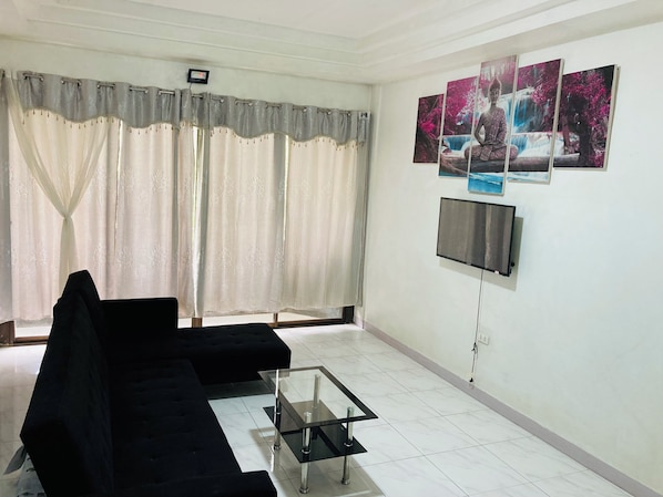A living room with a black sofa, a glass coffee table, and a flat-screen TV mounted on the wall.