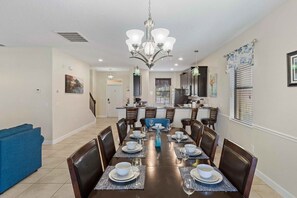 A dining area with a long table set for four, surrounded by chairs, with a kitchen in the background.
