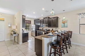 A modern kitchen with dark cabinets, a central island with bar stools, and a dining area with a window. - Resort Access 5BR Oasis w Pool, Game Room near WDW (Kissimmee)