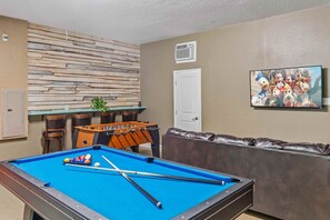 A pool table with pool cues and balls, a foosball table, a brown leather couch, and a wall-mounted television displaying animated characters. - Resort Access 5BR Oasis w Pool, Game Room near WDW (Kissimmee)