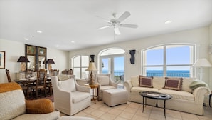 Uma sala de estar espaçosa com varanda vista para o mar, com ventilador de teto, janelas amplas e área de jantar com mesa e cadeiras.