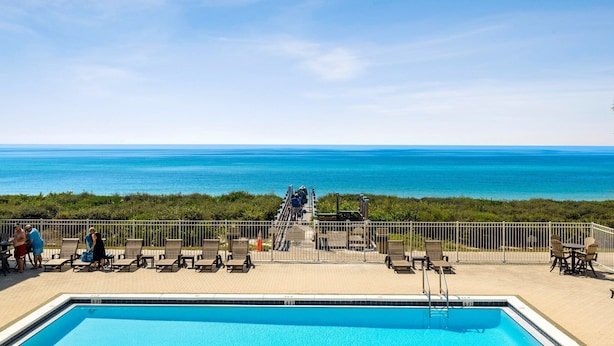 Uma área de piscina com vista para o oceano, um deck com cadeiras de sol e uma escadaria que dá para a praia.