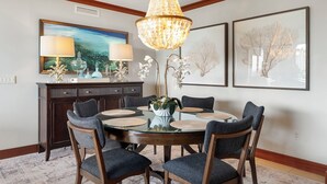 A dining area with a round table, six chairs, a chandelier, framed artwork, and a dark wooden cabinet.