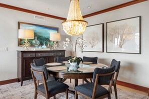 A dining area with a round table, six chairs, a chandelier, framed artwork, and a dark wooden cabinet.