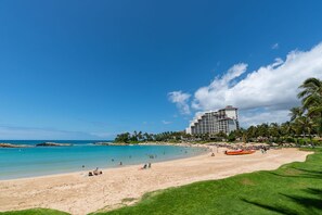 Una playa con un hotel al fondo, cielos despejados y palmeras.