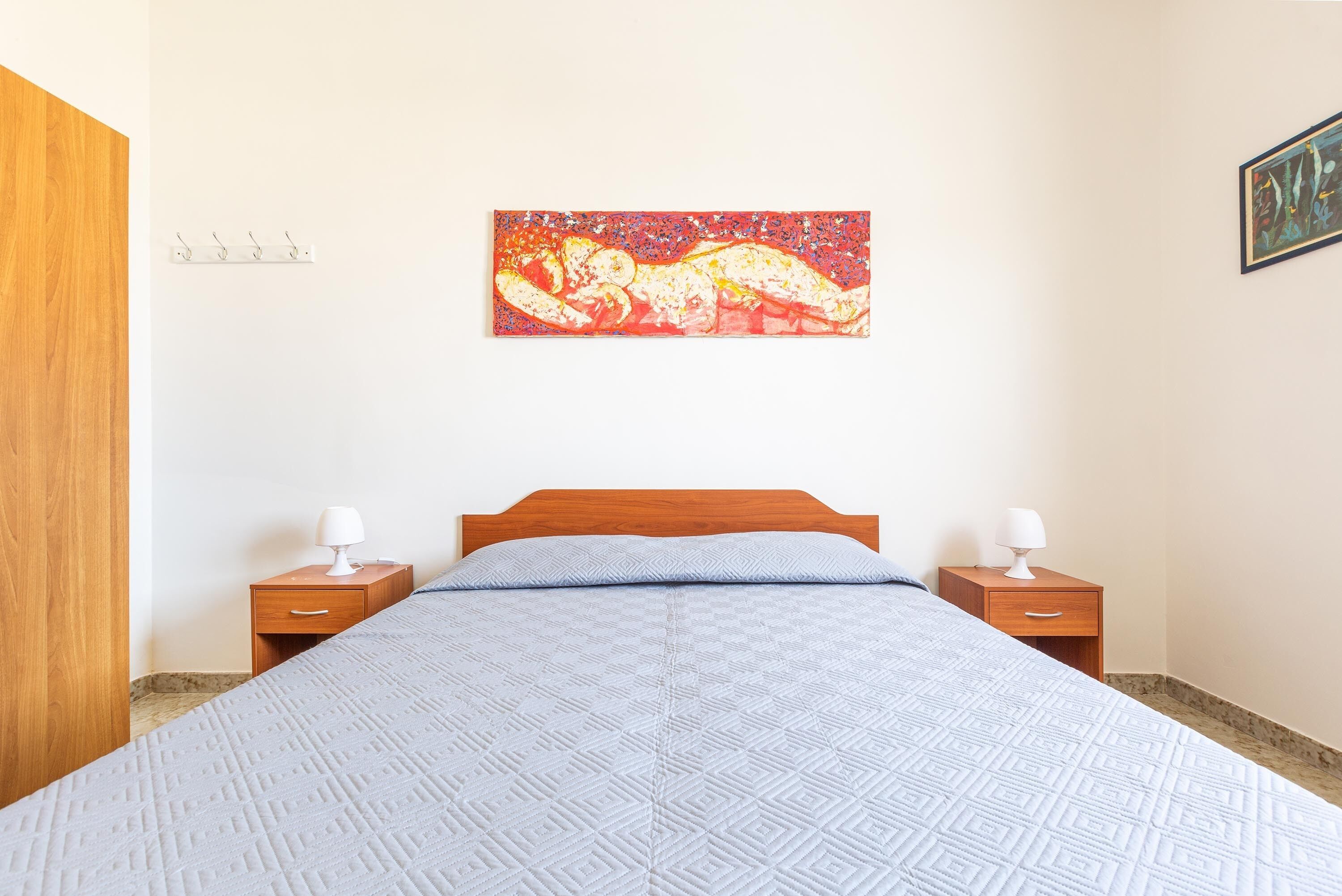 A bedroom with a bed, two bedside tables, a nightstand, a wall-mounted painting, and a wooden wardrobe.