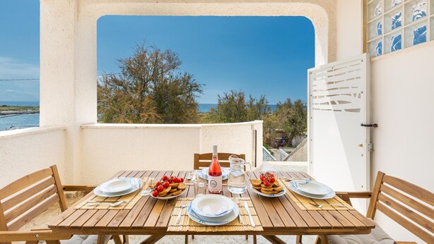 A wooden table set for two with plates, a bottle, and a pitcher, overlooking a scenic view of the sea and trees.