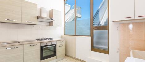 Una cocina moderna con alacenas de madera clara, estufa a gas y un ventanal con vistas a un edificio.