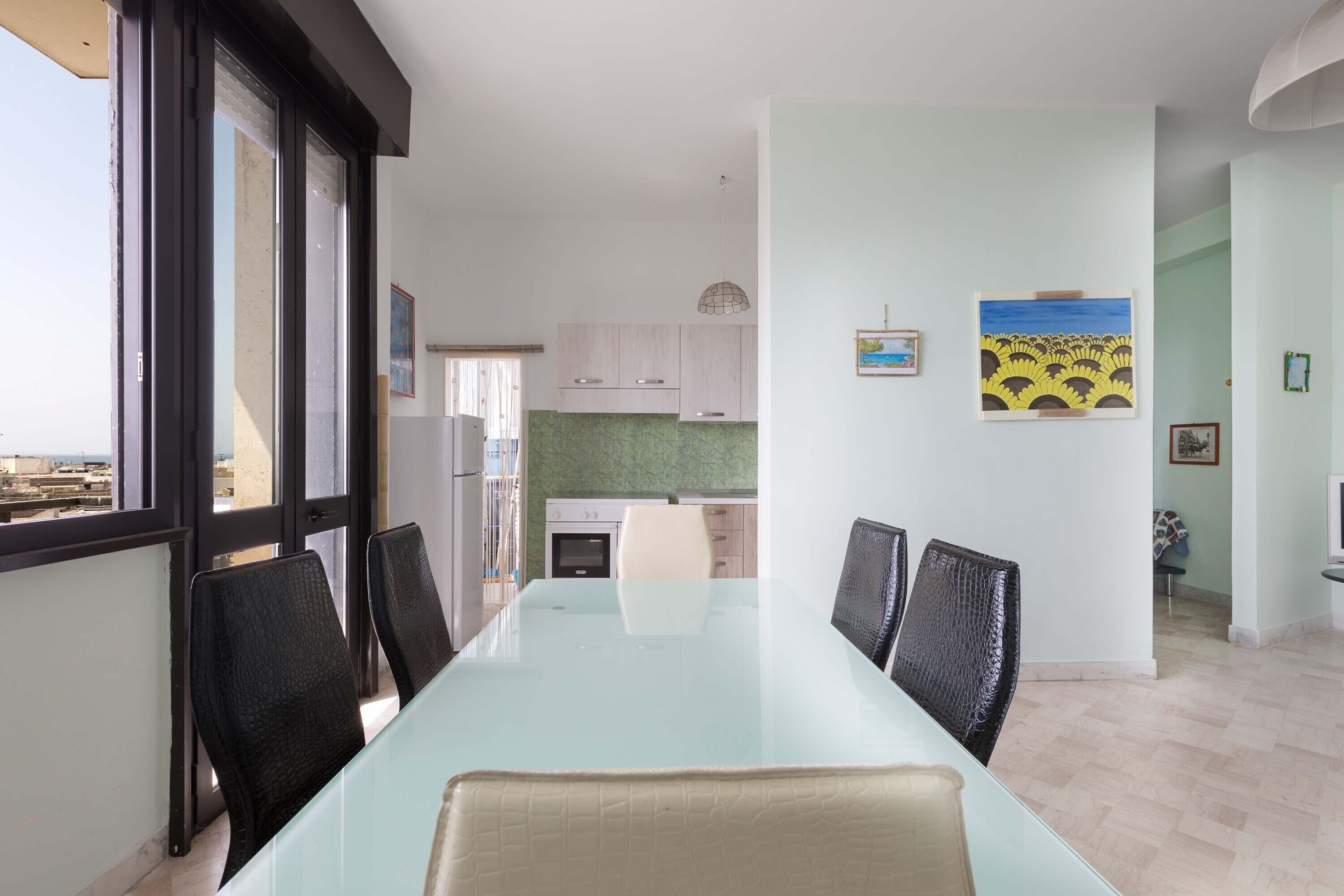 A dining area with a glass table and chairs, a kitchen in the background, and artwork on the walls.