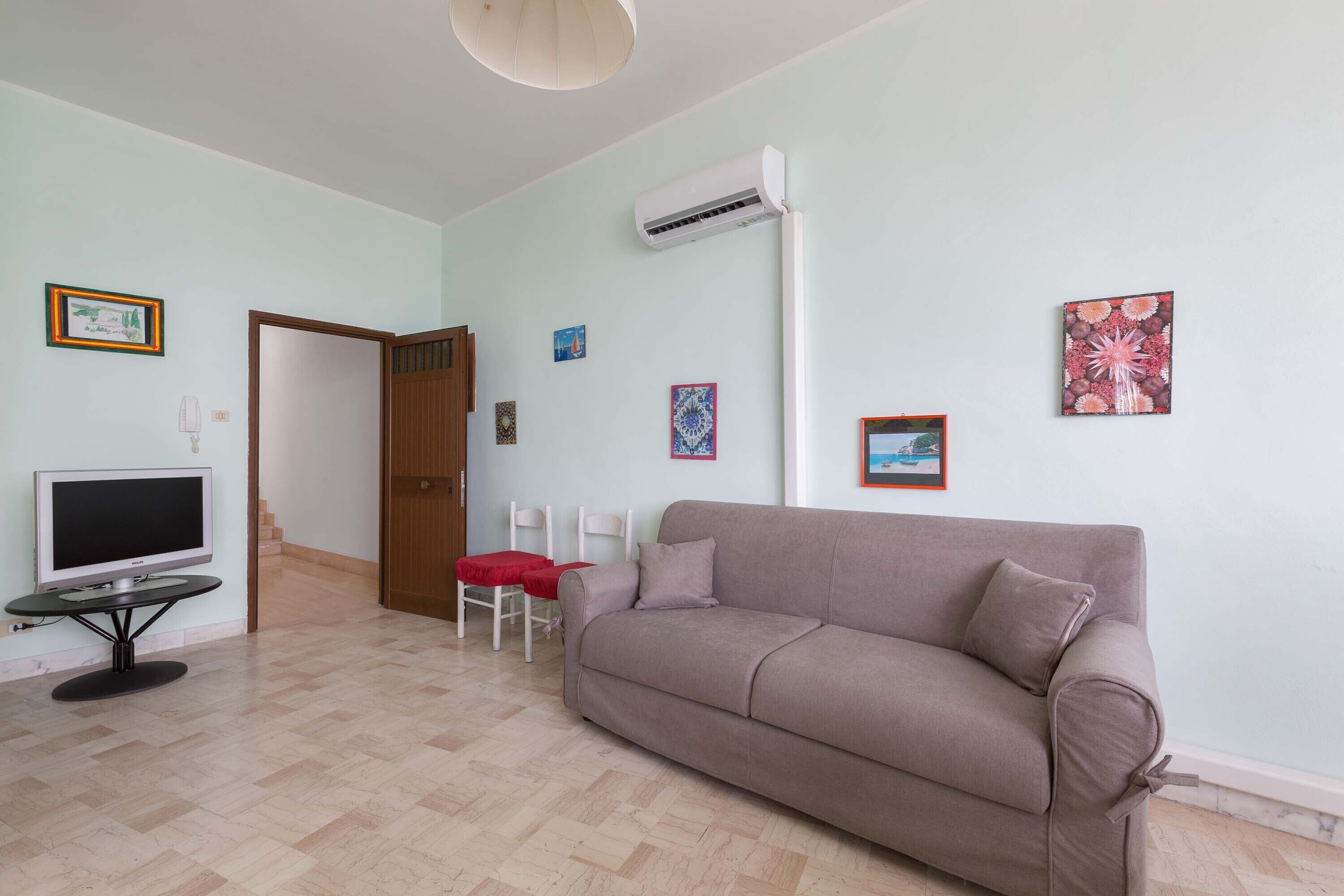 A living room with a beige sofa, a TV on a stand, a red table, and a door leading to another room.