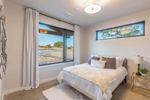 Een slaapkamer met een groot raam, een bed met een bloemenprint, een nachtkastje met een lamp, en een lijst met een afbeelding aan de muur.
