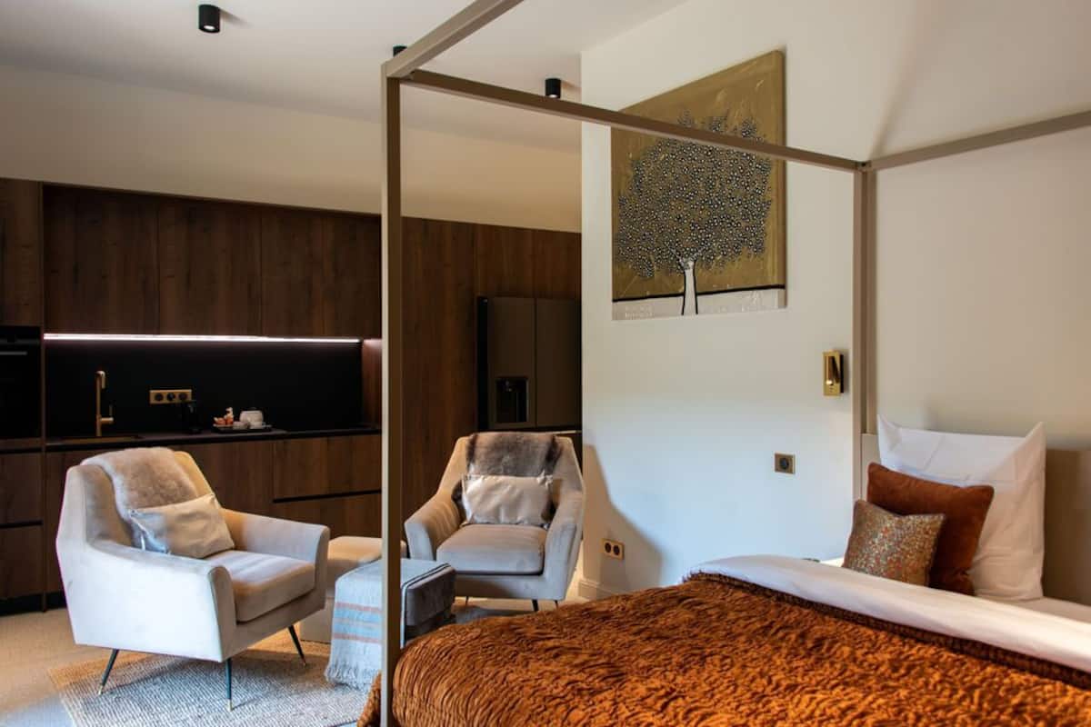 A modern hotel room with a large bed, two armchairs, a small table, and a large window.