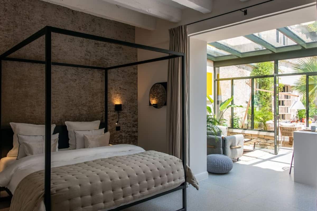 A four-poster bed with a tufted headboard, a stone-textured wall, and a view of a patio with outdoor furniture and plants.