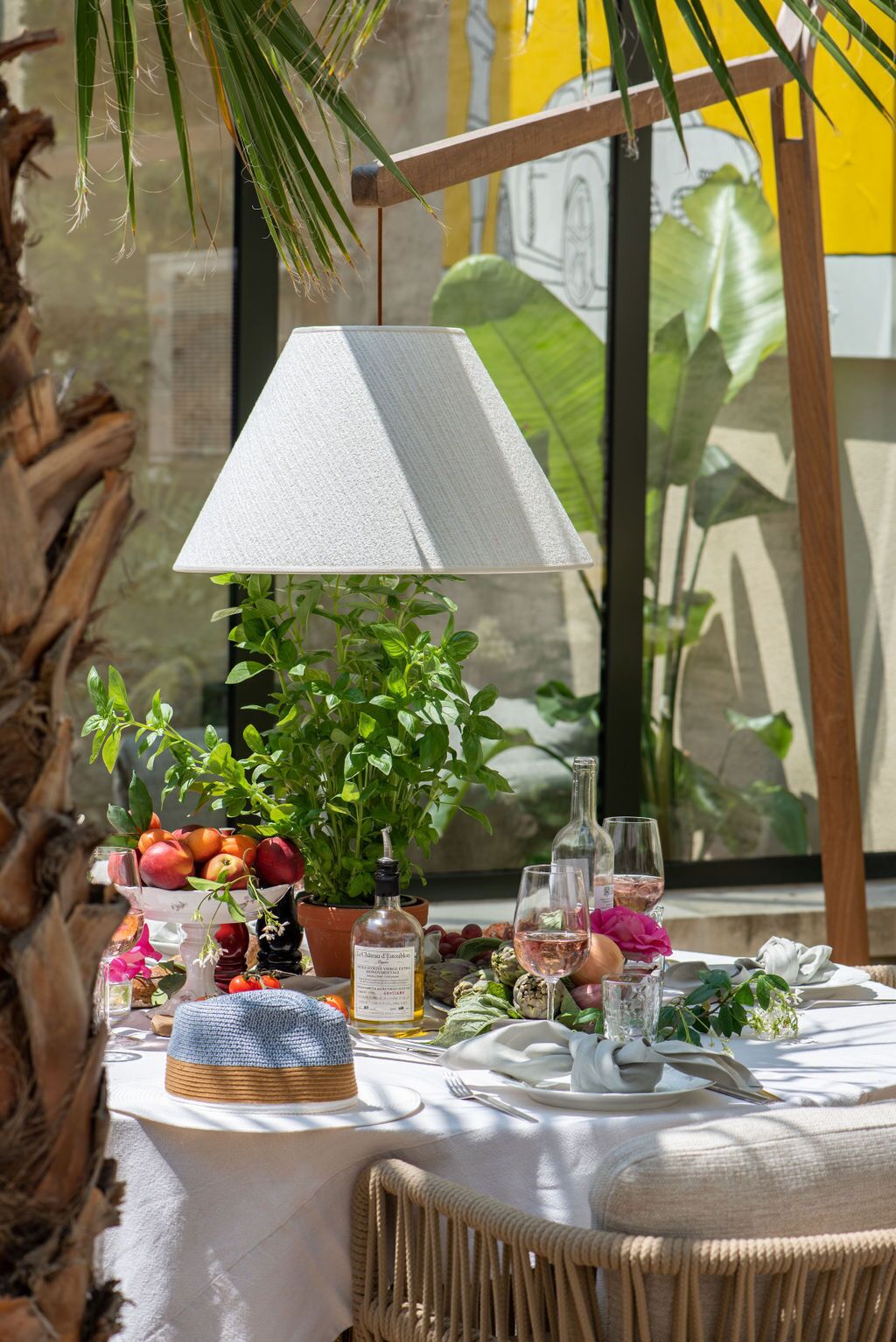 A table set with fruits, wine glasses, and a lamp in a bright, plant-filled space.