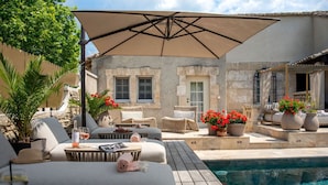 Zona junto a la piscina con mobiliario exterior, una pérgola y un edificio de piedra al fondo.