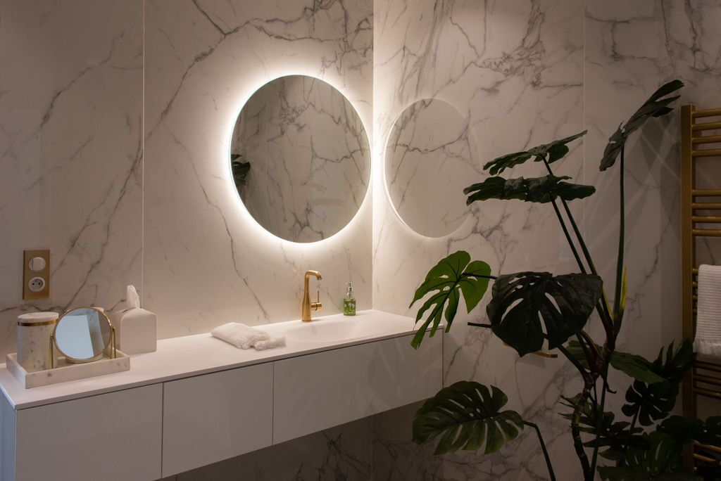 A modern bathroom with marble walls, a white vanity, a gold faucet, a round mirror, and a large plant.