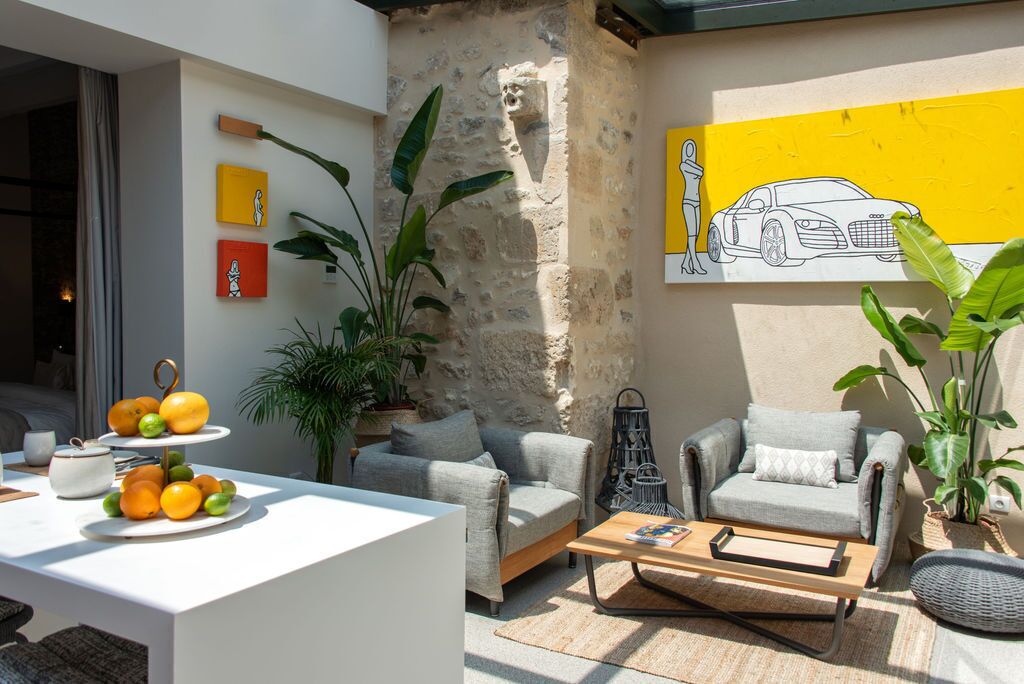 Un espace extérieur moderne avec des sièges, comprenant une table blanche, deux canapés gris et une table basse en bois. On y trouve également des plantes en pot et une œuvre d’art murale jaune.