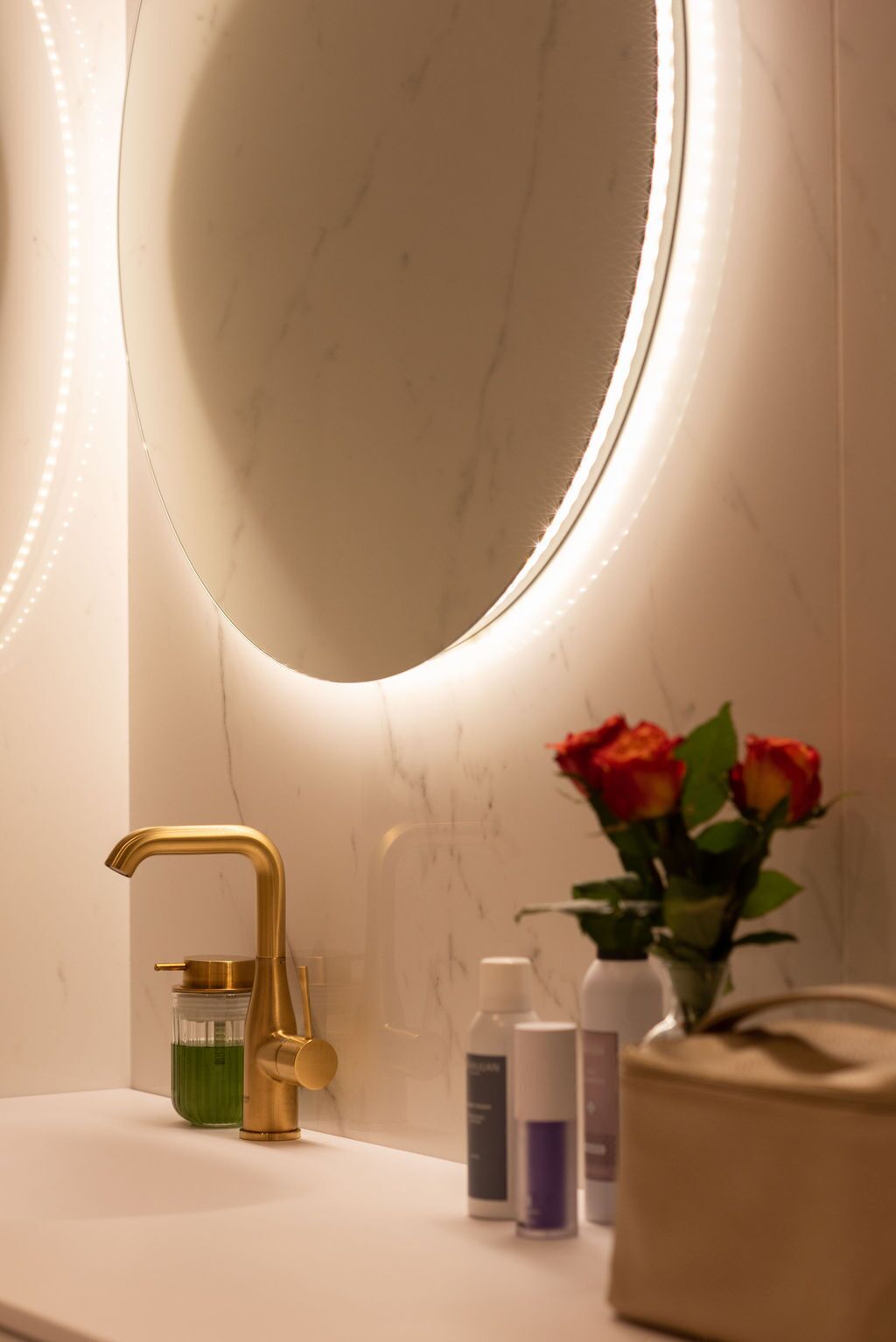 A bathroom with a gold faucet, a round mirror with LED lighting, and a vase with red roses on the counter.