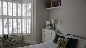 Une chambre à coucher comprenant un lit, une table de chevet avec une lampe, une chaise, un radiateur et une fenêtre avec des stores.