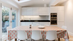Une cuisine moderne avec des meubles blancs, un plan de travail noir et des appareils en acier inoxydable. On y trouve une table à manger recouverte d’une nappe à motifs et entourée de chaises blanches.