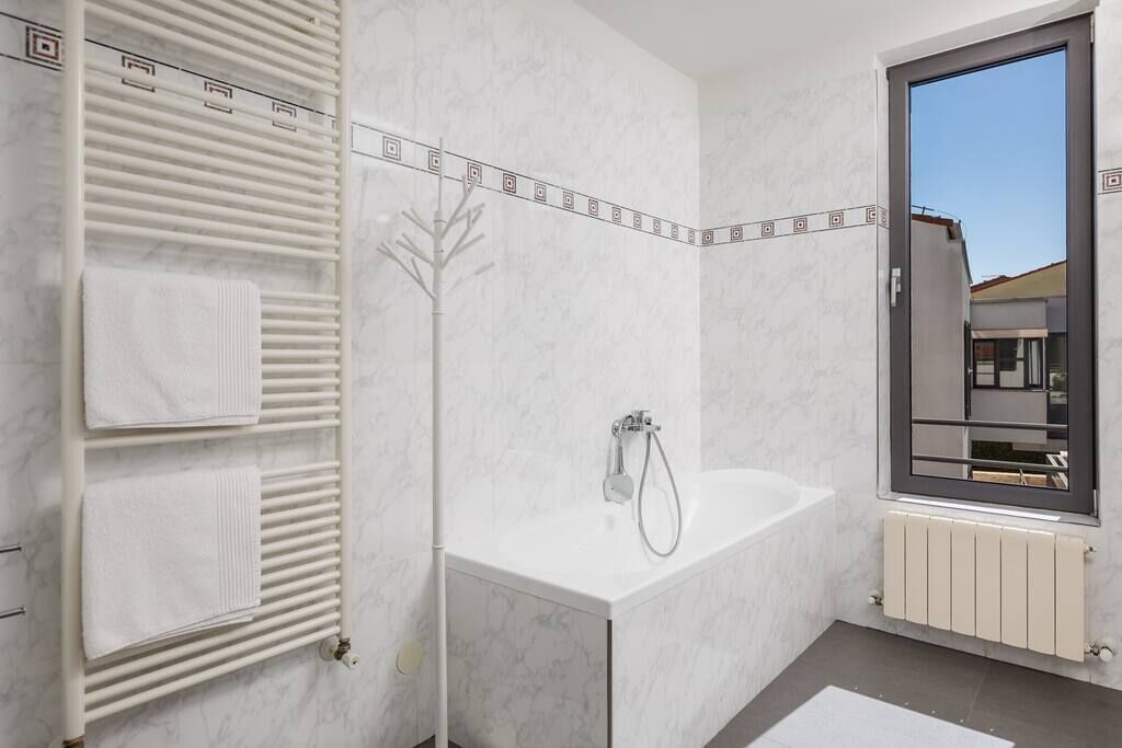 A modern bathroom with a white bathtub, a towel rack with folded towels, a radiator, and a window with a view of a building.