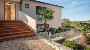Un bâtiment moderne avec un escalier menant à une porte en bois, une petite fenêtre et un jardin bien entretenu agrémenté de diverses plantes et d’un palmier.