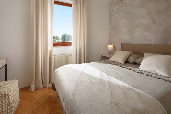 A bedroom with a large bed, a nightstand with a lamp, and a view of the outdoors through a window. - Villa Rosy in Rovinjsko Selo (Rovinjsko Selo)
