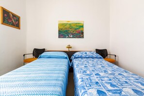 Una habitación con dos camas individuales, una mesita de noche con lámpara, un cuadro enmarcado en la pared y una silla con cojín naranja.